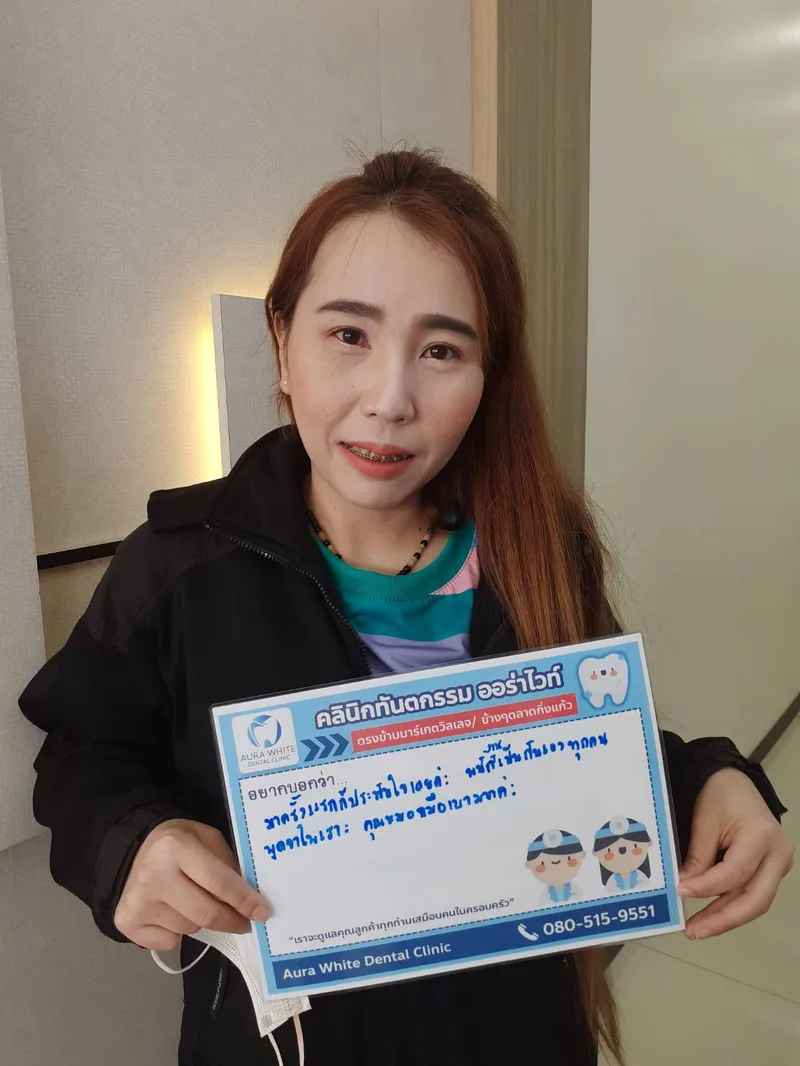 คนไข้หญิงติดเครื่องมือจัดฟันถือป้ายรีวิวชมราคาไม่แพงและการดูแลของทีมงาน คลินิกทันตกรรม AuraWhite บางพลี