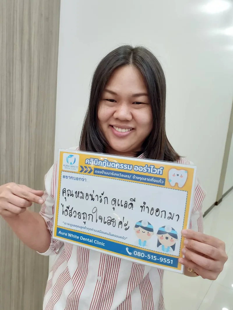 คนไข้หญิงถือป้ายรีวิวชมคุณหมอน่ารักดูแลดี ถ่ายในคลินิก คลินิกทันตกรรม AuraWhite บางพลี