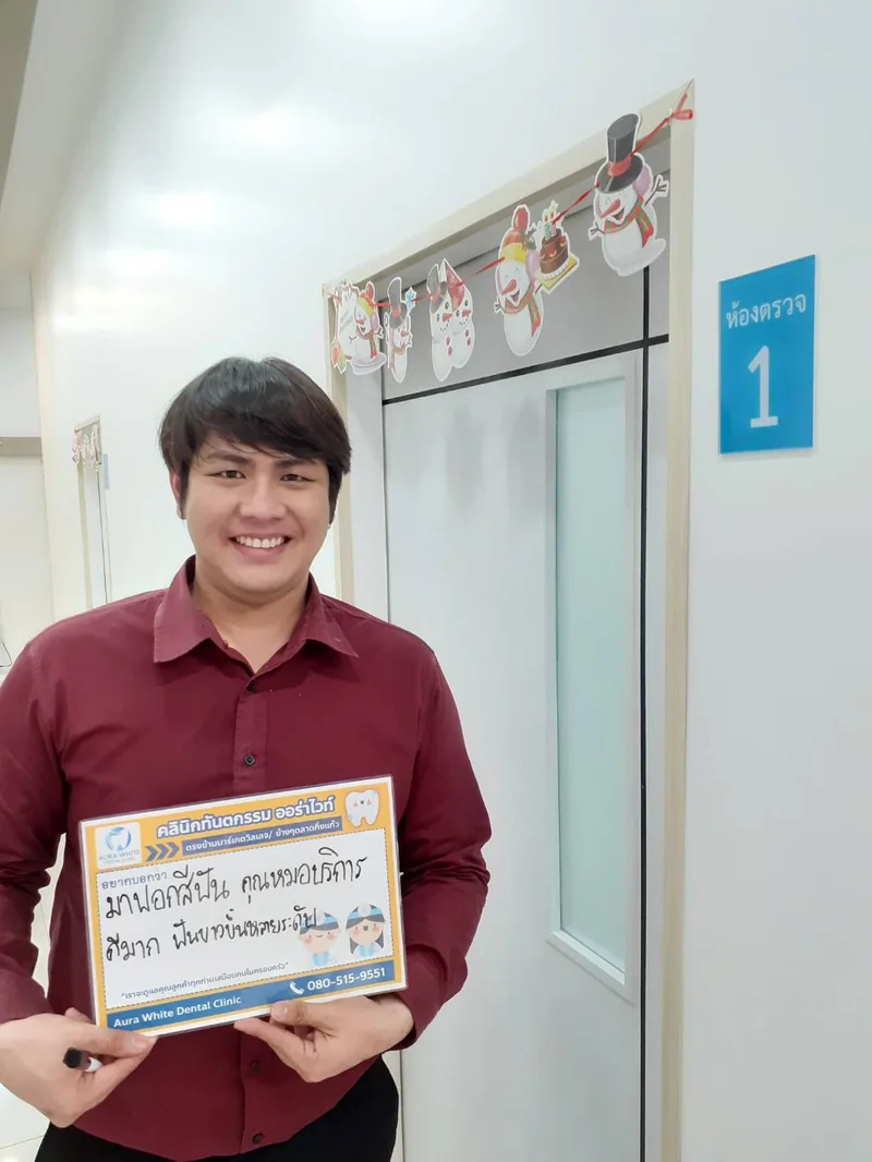 คนไข้ชายถือป้ายรีวิวชมบริการหลังฟอกสีฟัน หน้าห้องตรวจ 1 คลินิกทันตกรรม AuraWhite บางพลี
