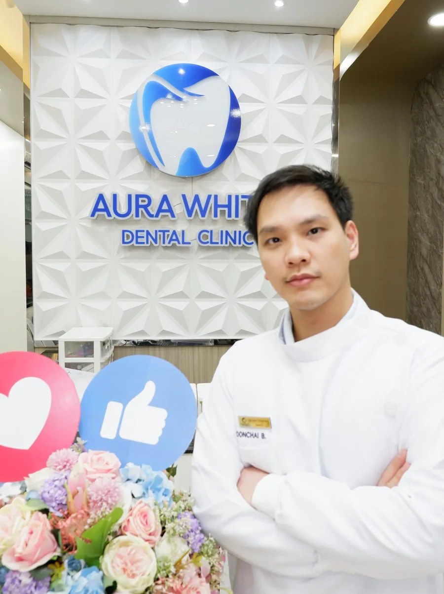 ทพ. บุญชัย บวรวราภรณ์ ยืนหน้าผนังโลโก้ Aura White Dental Clinic ภายในคลินิก